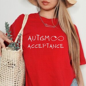 Może przedstawiać: Czerwony T-shirt z napisem "AUTISM ACCEPTANCE" w białym kolorze i symbolem nieskończoności.
