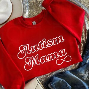 Może przedstawiać: Czerwony sweter z białym napisem "Autism Mama" w retro czcionce.