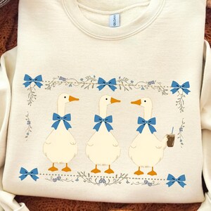 Könnte beinhalten: Cremefarbenes Sweatshirt mit drei Comic-Gänsen mit blauen Fliegen und orangefarbenen Schnäbeln. Die Gänse sind von einem floralen Design mit blauen Schleifen umgeben. Eine Gans hält ein braunes Getränk. Das Sweatshirt hat einen Rundhalsausschnitt.