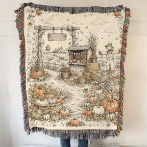 May include: A woven throw blanket featuring a detailed autumn harvest scene with pumpkins, a scarecrow, a wooden farm stand, and a rustic archway. The design is rendered in muted earth tones with a cream background and decorative fringed edges.