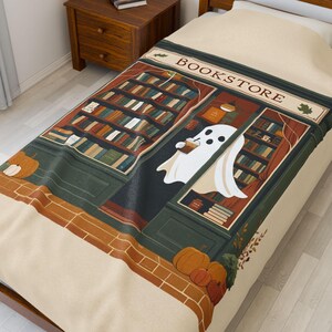 K&ouml;nnte beinhalten: Eine beige Decke mit einer Buchladen-Szene und einem Geist, der ein Getr&auml;nk h&auml;lt. Der Buchladen hat B&uuml;cherregale, und das Wort "BOOKSTORE" steht &uuml;ber dem Eingang. K&uuml;rbisse und Herbstlaub sind ebenfalls abgebildet.