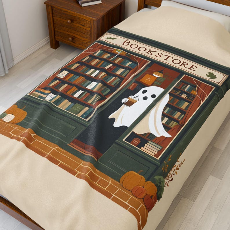 K&ouml;nnte beinhalten: Eine beige Decke mit einer Buchladen-Szene und einem Geist, der ein Getr&auml;nk h&auml;lt. Der Buchladen hat B&uuml;cherregale, und das Wort "BOOKSTORE" steht &uuml;ber dem Eingang. K&uuml;rbisse und Herbstlaub sind ebenfalls abgebildet.