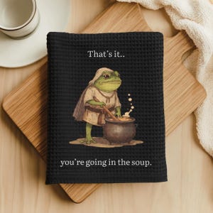 May include: A black waffle-weave kitchen towel featuring a graphic of a green frog wearing a hooded tunic and stirring a dark cooking pot. The text above the frog reads That's it.. and the text below the frog reads you're going in the soup.