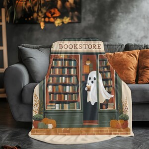 K&ouml;nnte beinhalten: Eine dekorative Decke mit einer Buchladen-Illustration. Das Design zeigt einen Geist mit einem Getr&auml;nk vor einem Buchladen, mit dem Wort "BOOKSTORE" &uuml;ber der T&uuml;r. K&uuml;rbisse und Pflanzen erg&auml;nzen das herbstliche Thema.