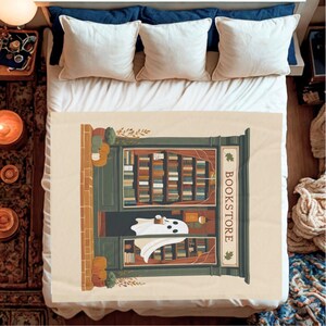 K&ouml;nnte beinhalten: Eine beige Decke mit einer Illustration eines Buchladens und einem Geist. Der Buchladen hat B&uuml;cherregale, einen gr&uuml;nen T&uuml;rrahmen und das Wort "BOOKSTORE". Der Geist h&auml;lt eine Tasse. Die Decke liegt auf einem Bett.