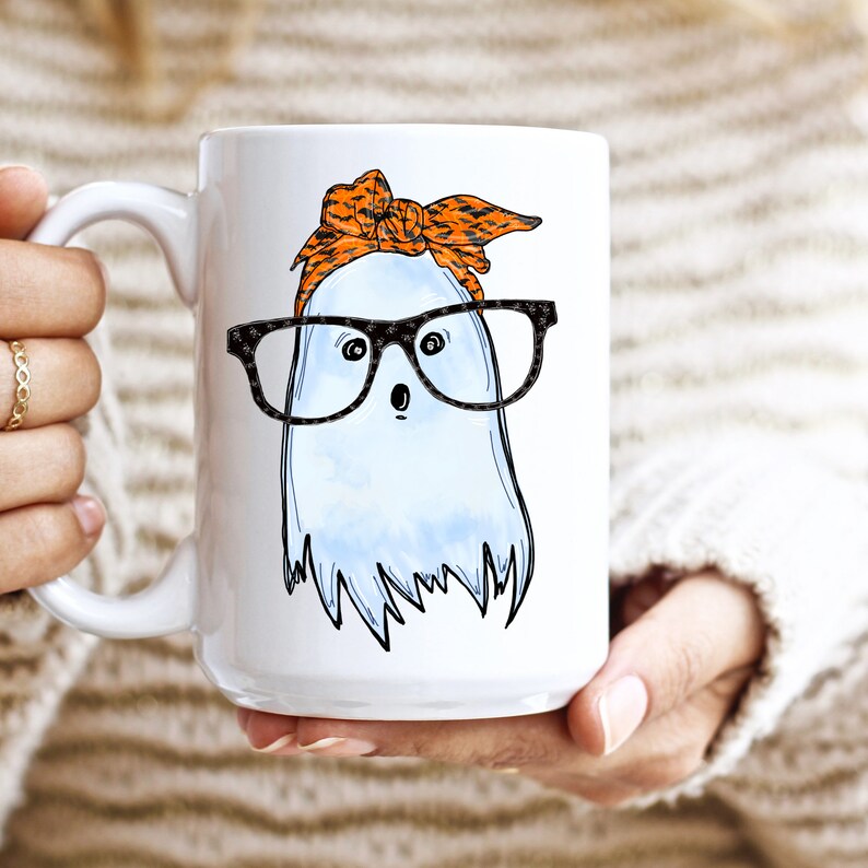 May include: White ceramic coffee mug with a cartoon ghost wearing orange and black patterned glasses and a headband. The ghost has a surprised expression.