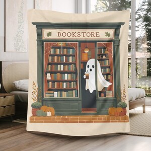 K&ouml;nnte beinhalten: Beige Decke mit einer Illustration eines Buchladens. Ein Geist mit einem Getr&auml;nk steht vor dem Buchladen. Das Wort "BOOKSTORE" steht &uuml;ber dem Eingang. K&uuml;rbisse und Pflanzen befinden sich am Fu&szlig;e des Ladens. Der T&uuml;rrahmen ist gr&uuml;n.