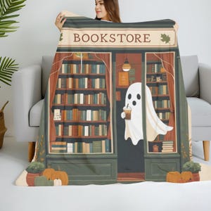 K&ouml;nnte beinhalten: Eine beige Decke mit einer Buchladen-Illustration. Das Design beinhaltet das Wort "BOOKSTORE", B&uuml;cherregale, K&uuml;rbisse und einen Geist, der ein Getr&auml;nk h&auml;lt. Die Decke ist &uuml;ber einem grauen Sofa drapiert.