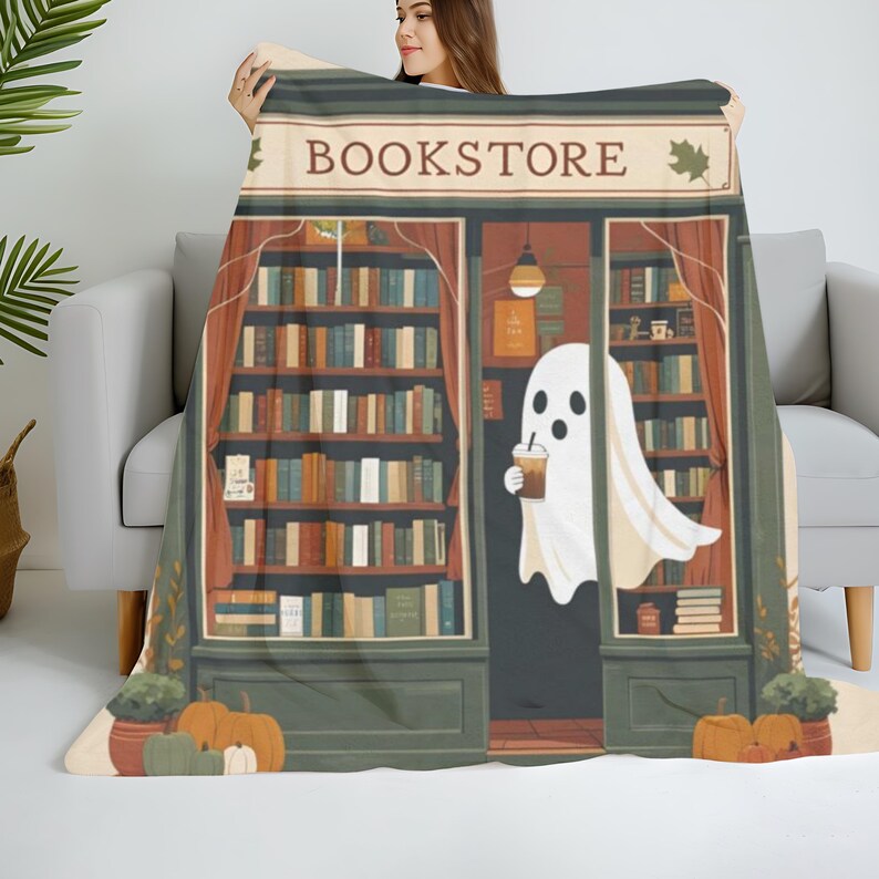 K&ouml;nnte beinhalten: Eine beige Decke mit einer Buchladen-Illustration. Das Design beinhaltet das Wort "BOOKSTORE", B&uuml;cherregale, K&uuml;rbisse und einen Geist, der ein Getr&auml;nk h&auml;lt. Die Decke ist &uuml;ber einem grauen Sofa drapiert.