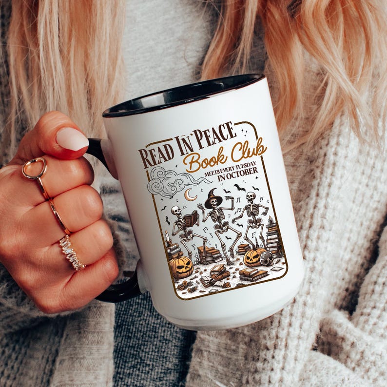 May include: White ceramic mug with a black interior and handle. The mug features the text "Read in Peace Book Club" with a Halloween-themed illustration of skeletons, pumpkins, and books. The mug is held by a person.