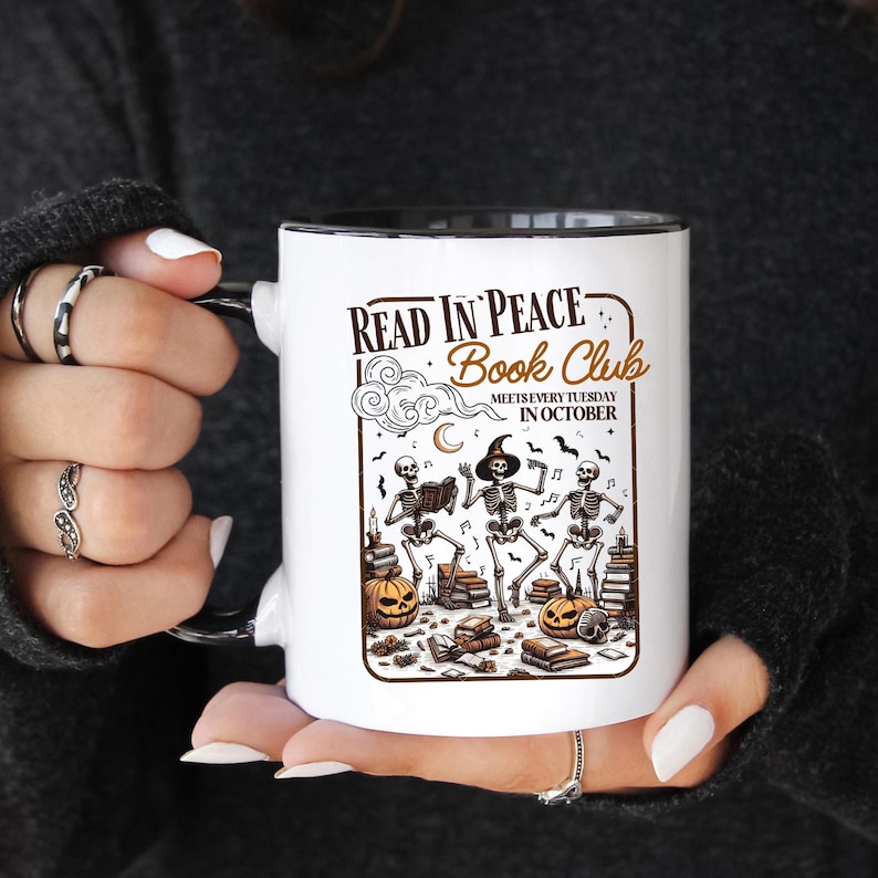 May include: White ceramic mug with a black interior and handle, featuring a Halloween-themed design. The design includes skeletons, pumpkins, books, and the text "Read in Peace Book Club". The mug is held by a person.