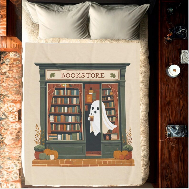 K&ouml;nnte beinhalten: Beige Decke mit einer Buchladen-Illustration. Die Szene zeigt eine gr&uuml;ne Ladenfront mit dem Wort "BOOKSTORE", einen Geist mit einem Getr&auml;nk und B&uuml;cherregale. K&uuml;rbisse und Pflanzen befinden sich am Fu&szlig;e des Ladens, rote Vorh&auml;nge sind zu sehen.
