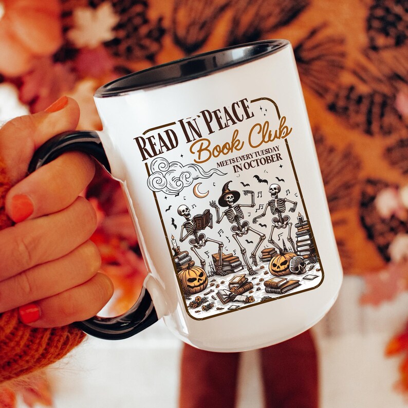 May include: White ceramic mug with a black handle and rim, featuring the text "READ IN PEACE Book Club" and a Halloween-themed illustration of skeletons, pumpkins, books, and a witch hat. The mug reads "MEETS EVERY TUESDAY IN OCTOBER".