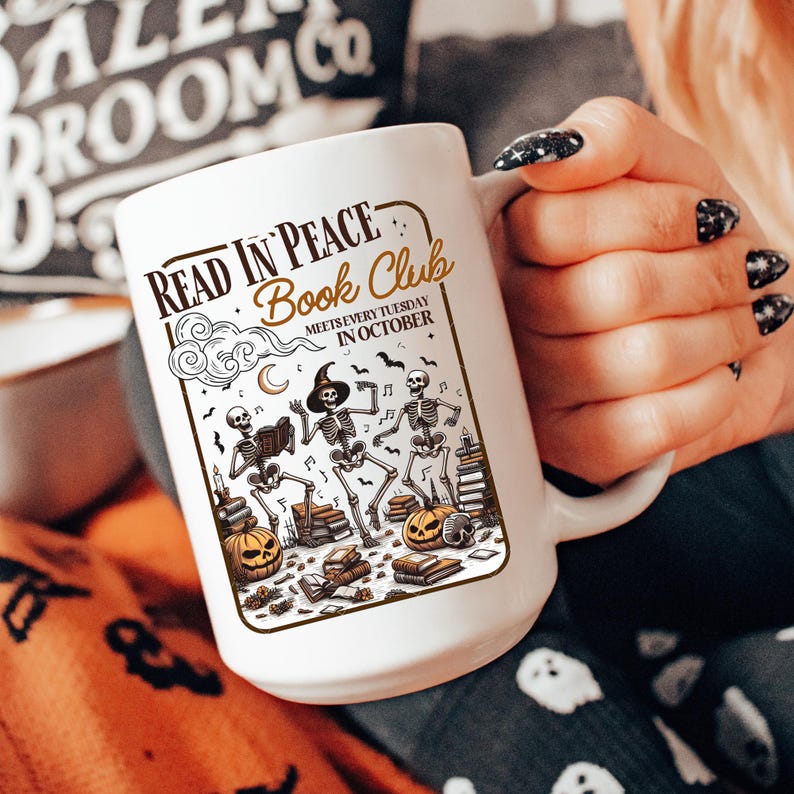 May include: White ceramic mug featuring a Halloween-themed design with skeletons, pumpkins, books, and the text "Read in Peace Book Club meets every Tuesday in October." The mug has a large handle and is held by a person.