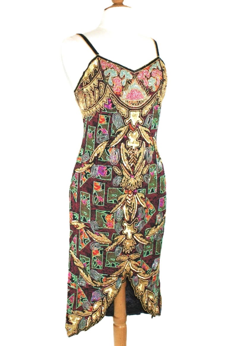 May include: A sleeveless midi dress in a deep burgundy hue, featuring an intricate pattern of geometric shapes and floral motifs. The garment is heavily embellished with gold, pink, blue, and green sequins and beads, with a V-neckline and thin straps.