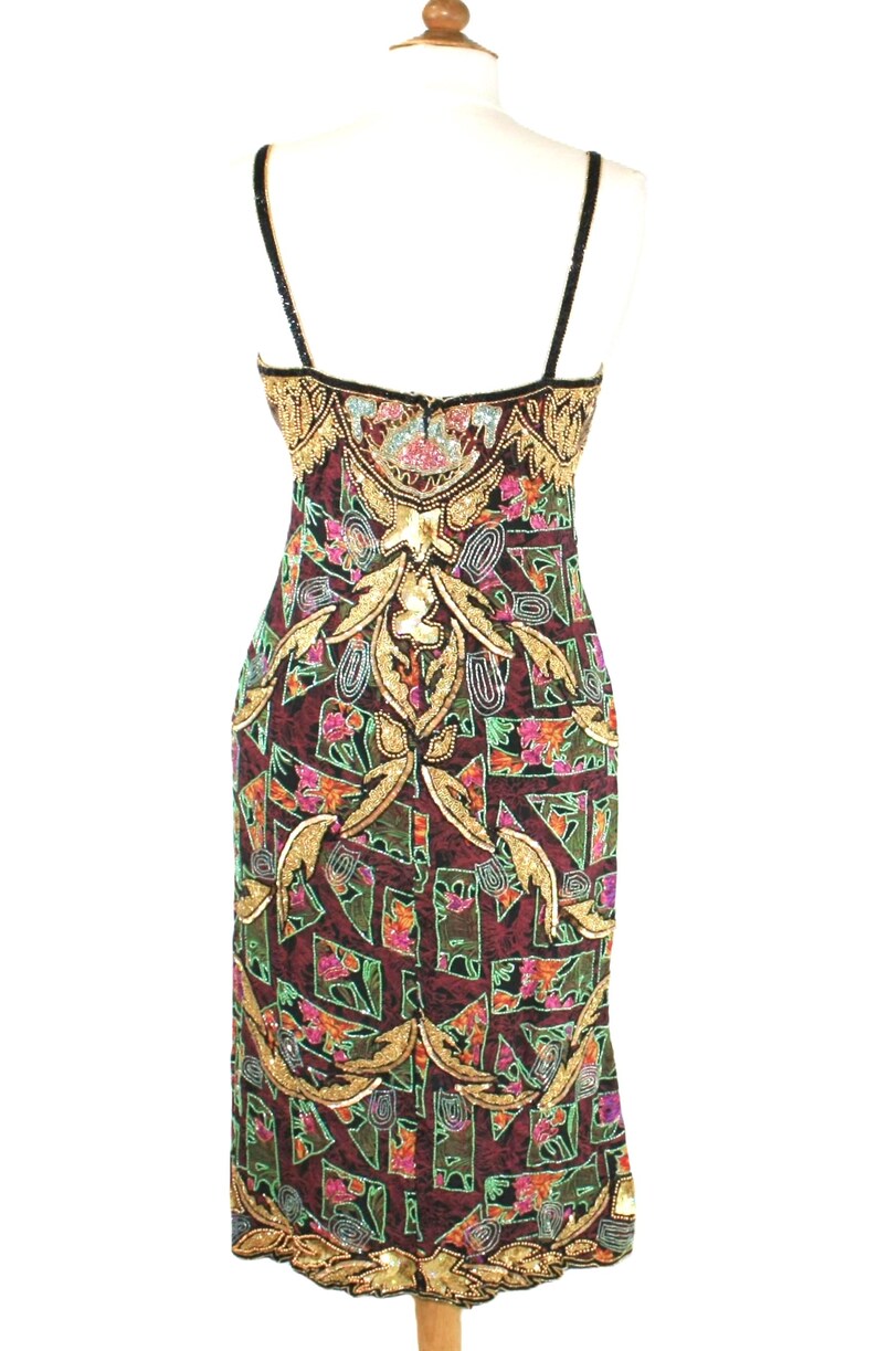 May include: A sleeveless sheath dress featuring a dark burgundy base with a geometric pattern in green, pink, and black. The garment is heavily embellished with gold sequins and beads in intricate floral and scroll motifs along the bodice and hem.