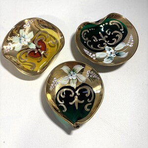 May include: Three small glass dishes with gold-toned rims and bases. Two dishes feature deep green centers with white floral patterns and gold scrollwork, while the third dish has a yellow center with similar floral and gold scroll designs.