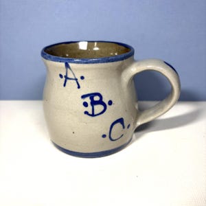 May include: A small, handmade ceramic mug with a light beige exterior and a dark brown interior. The rim and handle are accented with blue paint. The letters "A", "B", and "C" are hand-painted in blue on the front of the mug.