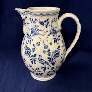 May include: A white ceramic pitcher with a blue floral pattern and a bird design. The pitcher has a curved handle and a spout for pouring. The design includes flowers, leaves, and vines. The pitcher is a decorative item.
