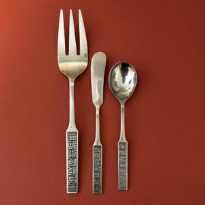 May include: A set of three silver-toned utensils with geometric patterns on the handles. The set includes a fork, a butter knife, and a spoon.