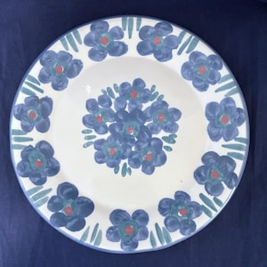 May include: A round, white ceramic plate with a blue floral design. The plate features a ring of blue flowers with red centers and green accents around the rim. The plate's edge has a blue border.