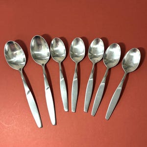 May include: Seven stainless steel spoons arranged on a red surface. Each spoon has a rounded bowl and a long, slender handle. The spoons are evenly spaced, reflecting light and creating a clean, modern aesthetic. Ideal for serving or eating.