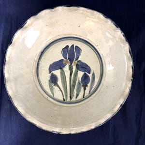 May include: A handmade ceramic plate with a cream-colored base and a blue iris floral design. The plate has a scalloped edge and a blue and green border around the floral center. The iris is painted in shades of blue and green.