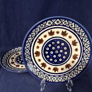 May include: Two round ceramic plates featuring a central dark blue circle decorated with small white and green dots. A cream-colored band surrounds the center, adorned with brown maple leaf motifs, finished with a blue and white lattice border.