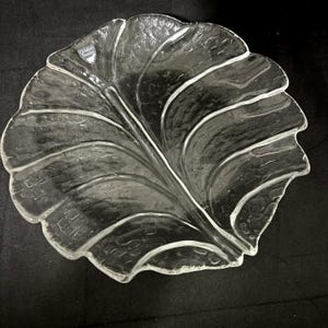 May include: Clear glass leaf-shaped serving dish. The dish has a textured surface and a detailed leaf vein design. The dish is made by Orrefors Sweden. The dish is perfect for serving snacks or as a decorative piece.