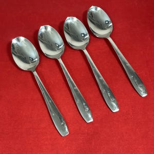 May include: A set of four polished silver-tone metal spoons arranged in a diagonal row. Each spoon features a smooth, rounded bowl and a flat handle decorated with a series of small, engraved star-shaped patterns along the lower portion of the stem.