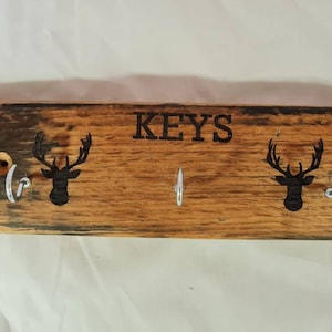 Könnte beinhalten: Schlüsselhalter aus Holz mit dem Wort "KEYS" und zwei Hirschkopf-Silhouetten. Der Schlüsselhalter hat drei silberne Haken zum Aufhängen von Schlüsseln. Das Holz hat ein natürliches, rustikales Aussehen.
