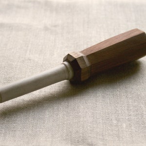 May include: A wooden knife sharpener with a dark brown, faceted handle and a light gray, cylindrical sharpening rod. The tool is designed for sharpening knives and other cutting tools. The sharpener is approximately 7 inches long.