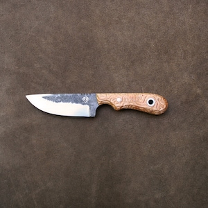 Puede incluir: Un pequeño cuchillo hecho a mano con un mango de madera marrón y una hoja negra. La hoja tiene una ligera curva y una punta puntiaguda. El mango tiene un patrón único en espiral y un pequeño anillo de metal cerca del extremo.