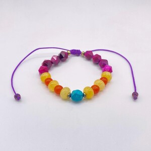 May include: A colorful beaded bracelet featuring a variety of gemstone beads in shades of purple, pink, yellow, orange, and turquoise. The bracelet is strung on a purple cord with adjustable ends.