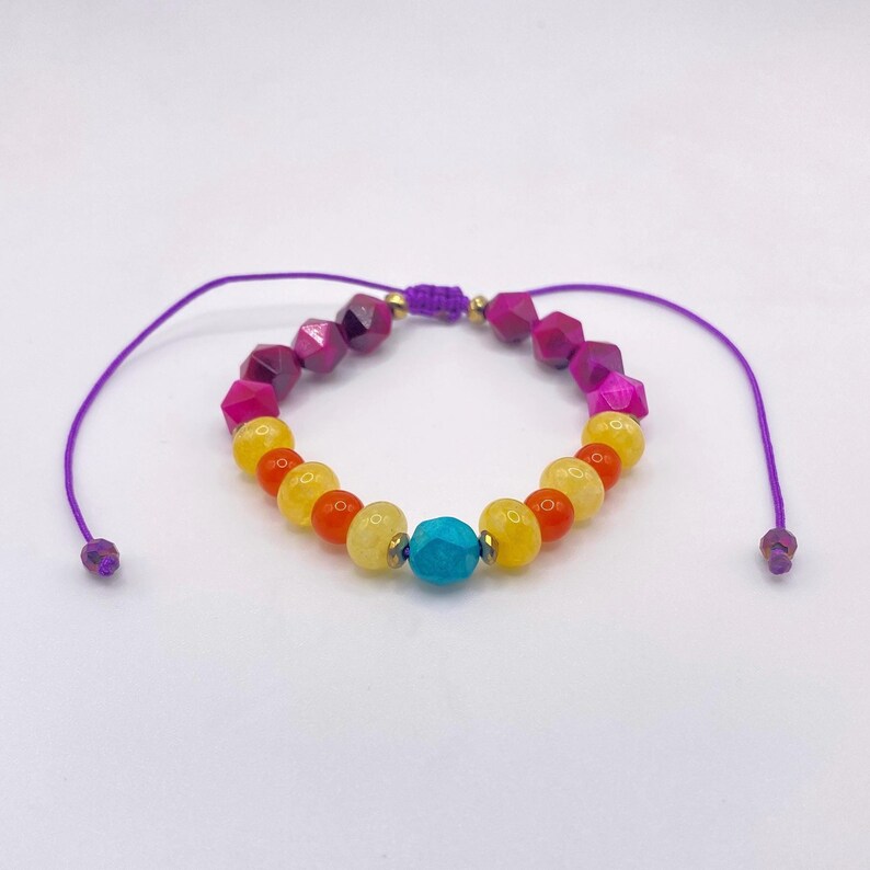 May include: A colorful beaded bracelet featuring a variety of gemstone beads in shades of purple, pink, yellow, orange, and turquoise. The bracelet is strung on a purple cord with adjustable ends.