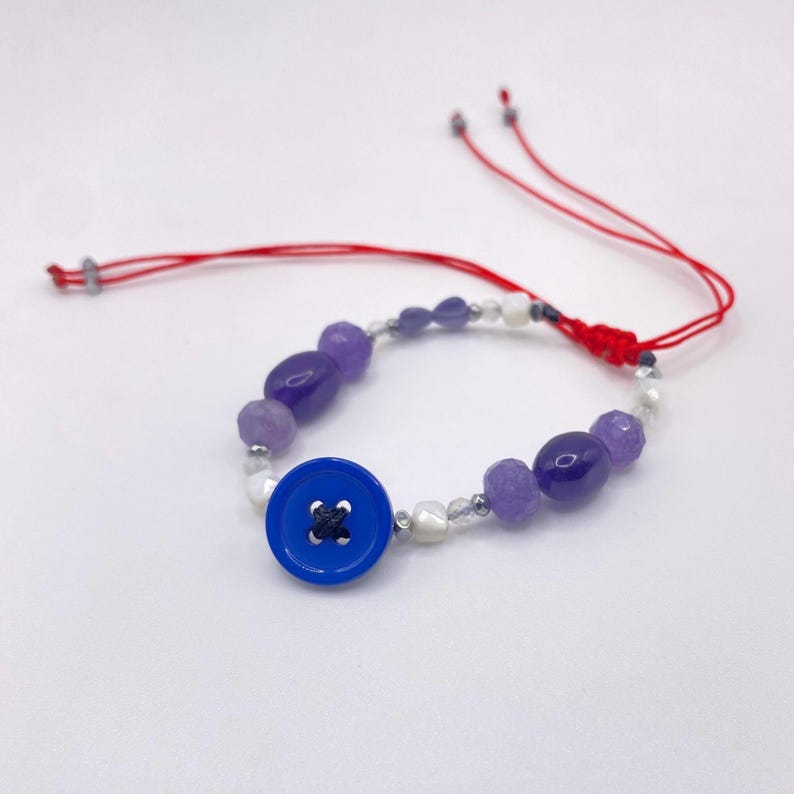 May include: A handmade bracelet featuring a vibrant blue button as a focal point, complemented by purple and white beads. The bracelet is strung on a red cord, creating a striking contrast against the white background.
