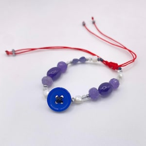 May include: A handcrafted bracelet with a prominent blue button, complemented by purple and white beads. The bracelet is strung on a red cord, offering a distinctive and vibrant accessory.
