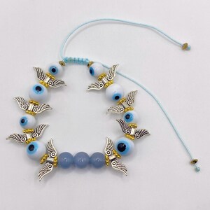 May include: A light blue adjustable bracelet featuring white and blue evil eye beads, silver-colored wing charms, and light blue beads. The bracelet is strung on a light blue cord with adjustable knots.