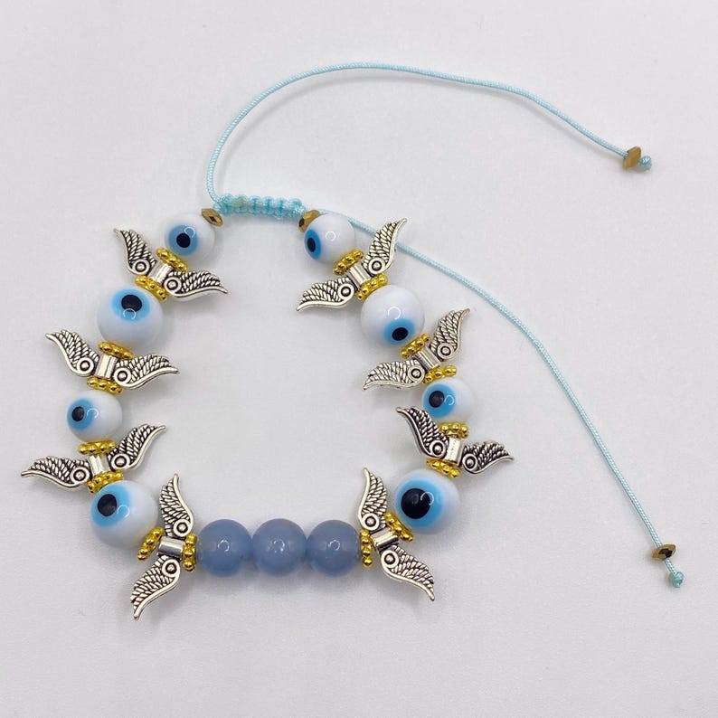 May include: A light blue adjustable bracelet featuring white and blue evil eye beads, silver-colored wing charms, and light blue beads. The bracelet is strung on a light blue cord with adjustable knots.
