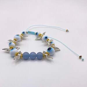 May include: A blue and white beaded bracelet featuring multiple evil eye charms with silver-colored wings and gold accents. The bracelet also includes blue beads and a light blue adjustable cord.