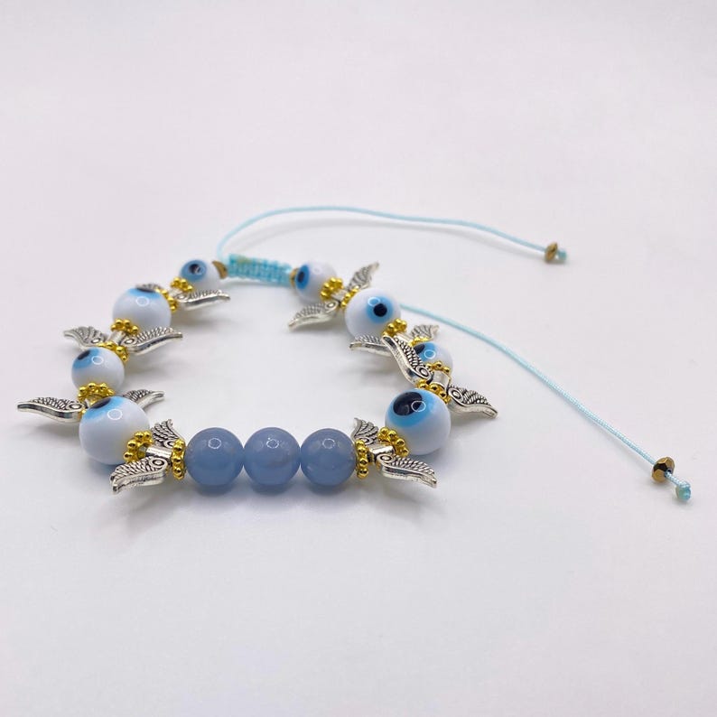 May include: A blue and white beaded bracelet featuring multiple evil eye charms with silver-colored wings and gold accents. The bracelet also includes blue beads and a light blue adjustable cord.