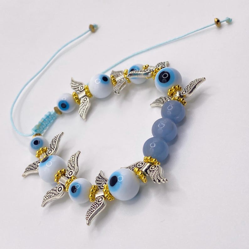 May include: A light blue adjustable bracelet featuring a series of white and blue evil eye beads, interspersed with silver-colored wing charms and gold accents. The bracelet also includes blue-grey beads.