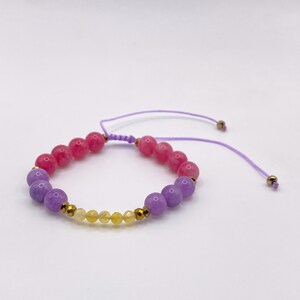 May include: A beaded bracelet featuring pink, purple, and yellow gemstones. The bracelet has a sliding knot closure with a purple cord and gold-colored accents. The beads are round and vary in size, creating a colorful accessory.