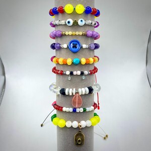 May include: A collection of beaded bracelets in various colors and designs. The bracelets feature a variety of beads, including colorful glass, gemstones, and unique charms. The bracelets are displayed on a gray cylindrical stand.