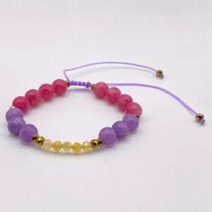 May include: A bracelet with pink, purple, and yellow beads. The bracelet has a sliding knot closure with a purple cord. Gold-coloured beads are interspersed among the yellow beads.