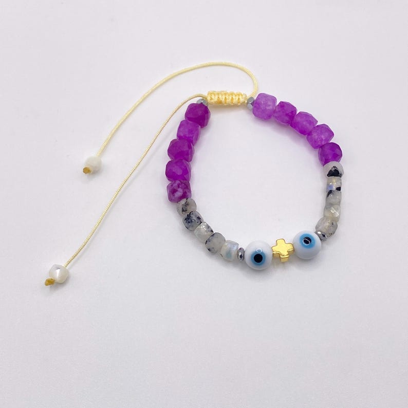 May include: Adjustable bracelet featuring purple and gray gemstone beads. The bracelet includes two blue evil eye beads and a gold-colored cross charm. The bracelet is strung on a cream-colored cord.