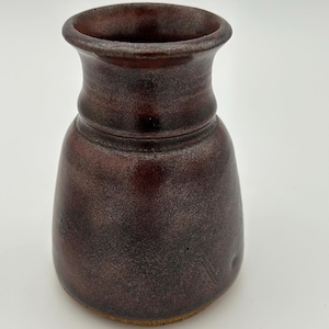 Peut inclure: Un vase en céramique brun foncé fait à la main, avec un bord évasé et un corps arrondi. Le vase a une surface texturée et un aspect moucheté, ajoutant à son charme rustique. Le vase mesure environ 15 cm de haut.