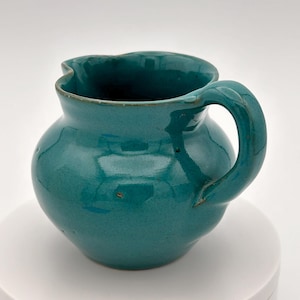 Peut inclure: Un petit pichet en céramique bleu turquoise avec un corps arrondi et un bord légèrement évasé. Le pichet a une anse qui se courbe vers le haut et au-dessus du bord.