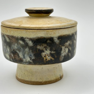 May include: A ceramic lidded jar with a pedestal base. The jar has a brown and white glaze with a mottled pattern. The lid is a light brown colour.