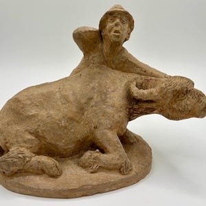 May include: A terracotta sculpture of a man holding onto a buffalo. The man is wearing a hat and has a distressed expression on his face. The buffalo is lying down with its head turned to the side.